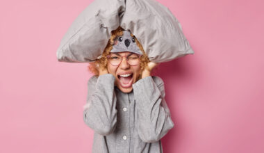 Horizontal shot of emotional woman exclaims loudly cannot fall asleep because of noise keeps soft pillow over head covers ears dressed in nightwear isolated over pink background. Sleep interruption